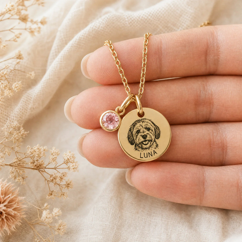 Hand holding the custom pet portrait necklace showing the engraved Havanese face, the name LUNA, and a pink birthstone charm on dried flowers and linen.
